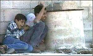 Photo shows a young Palestinian boy crying in terror as he huddles against his father, as both press against a wall next to a cement structure protruding from the wall. The father is waving to somebody and looking around the edge of the cement structure as he calls out.